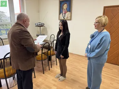 Mężczyzna wręcza dokument młodej kobiecie, obok stoi druga kobieta w sali.